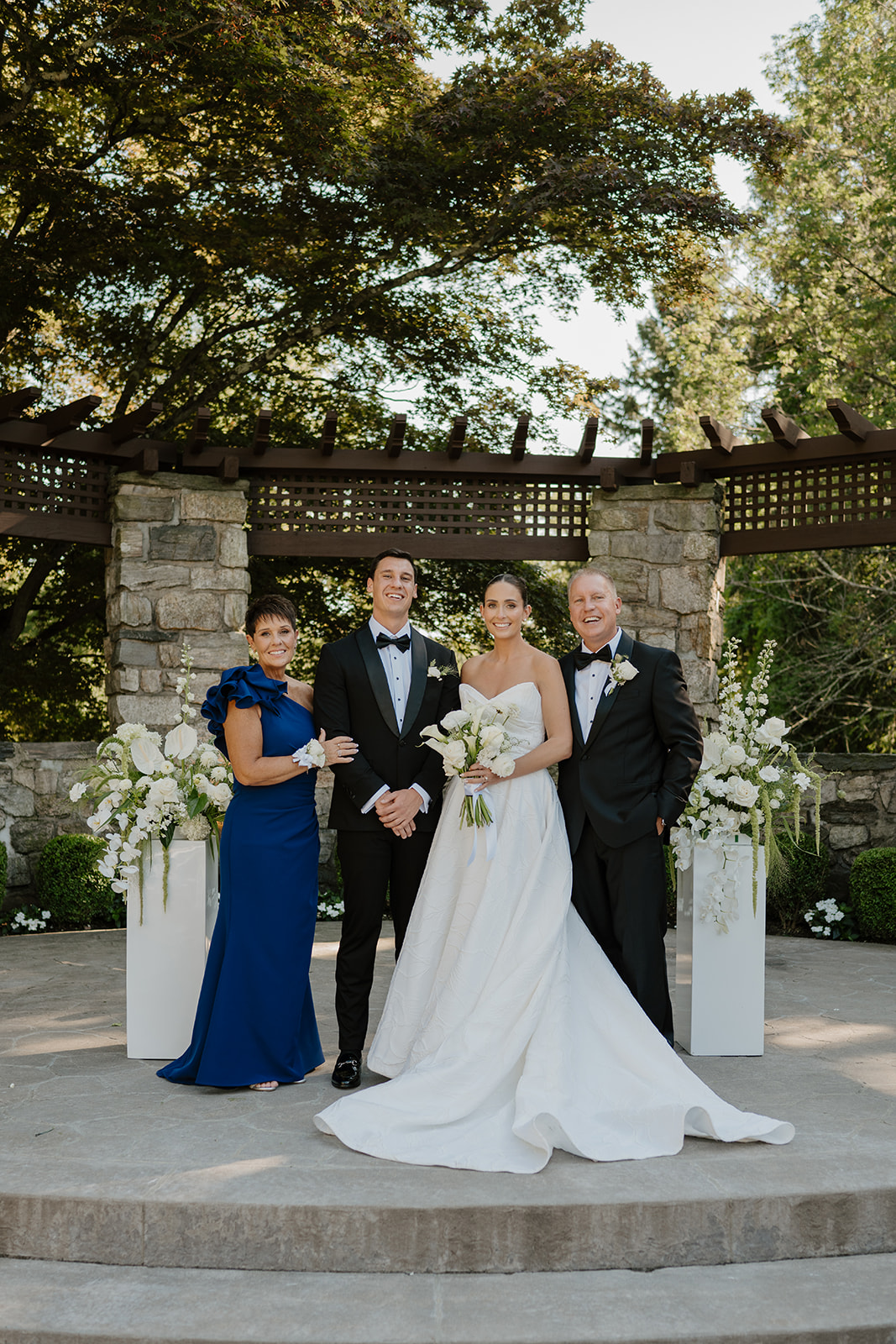 Bride and groom smiling with the groom's parents under a wooden arbor decorated with white flowers.